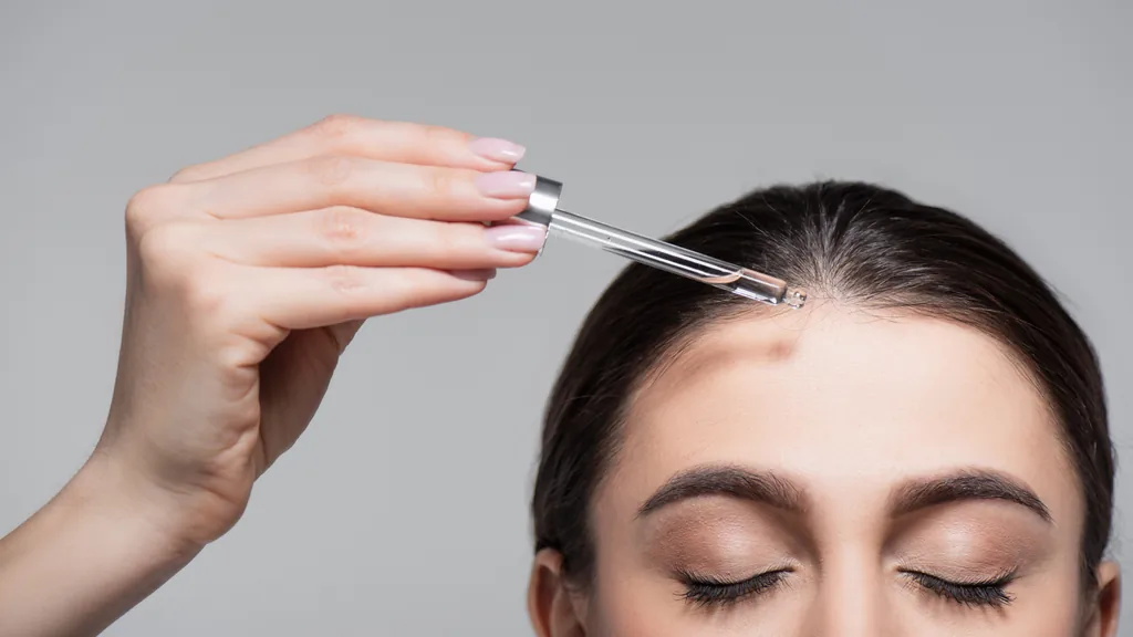Woman applying a clear dropper serum along her hairline with a pink manicure